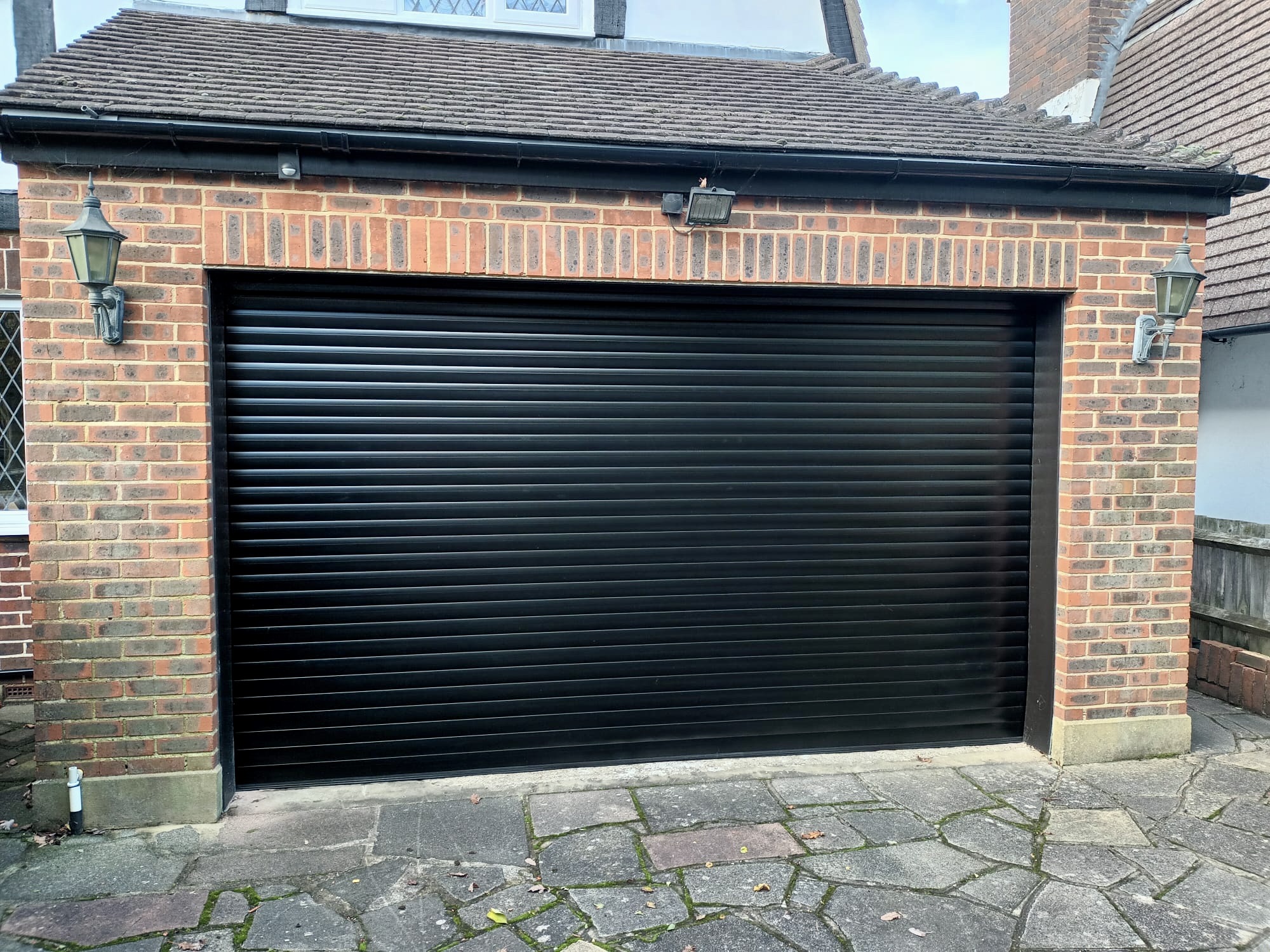 A black roller garage door set within a brick structure, flanked by wall lights.