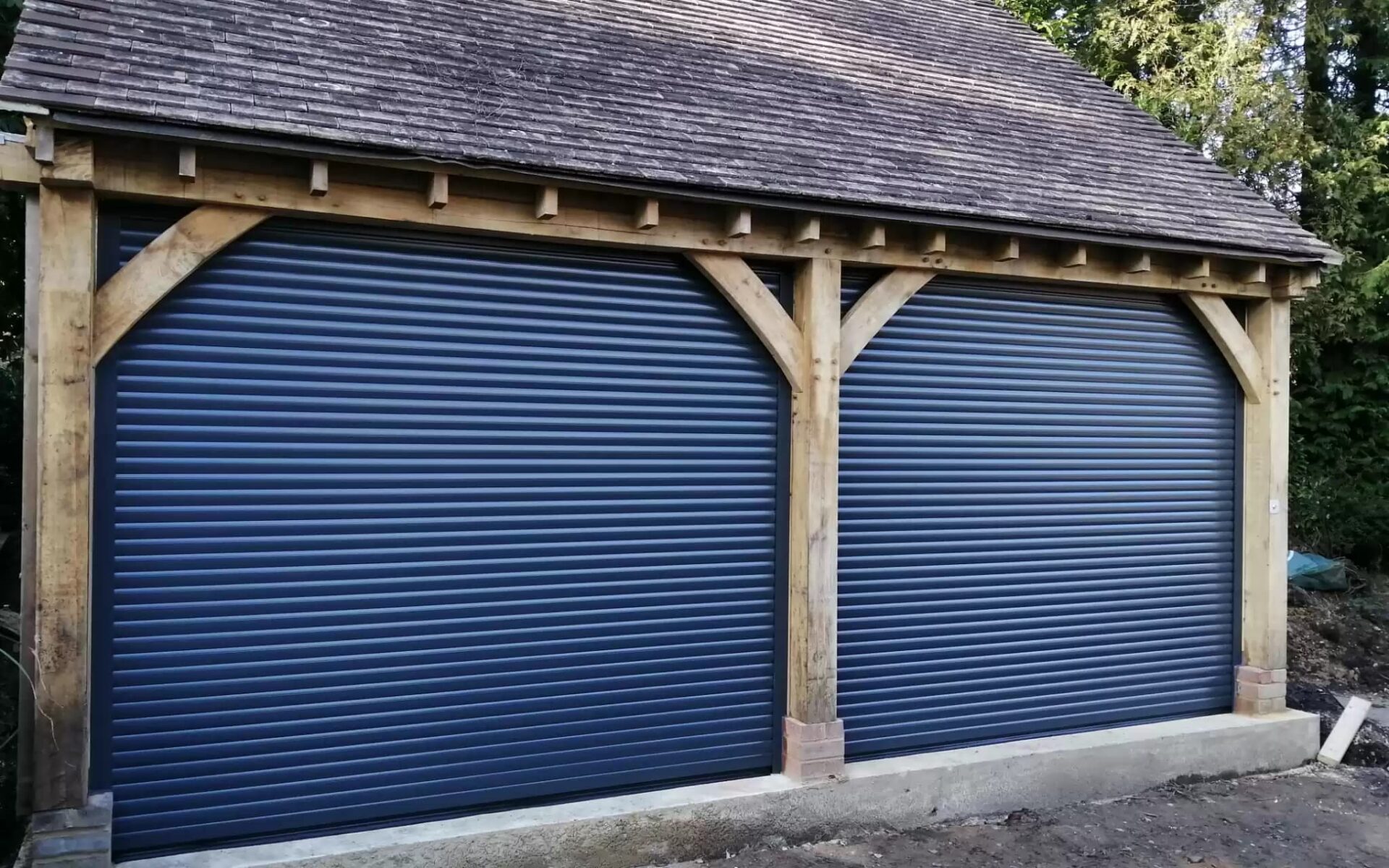 Two dark blue roller garage doors installed in a wooden-framed garage structure with a tiled roof.