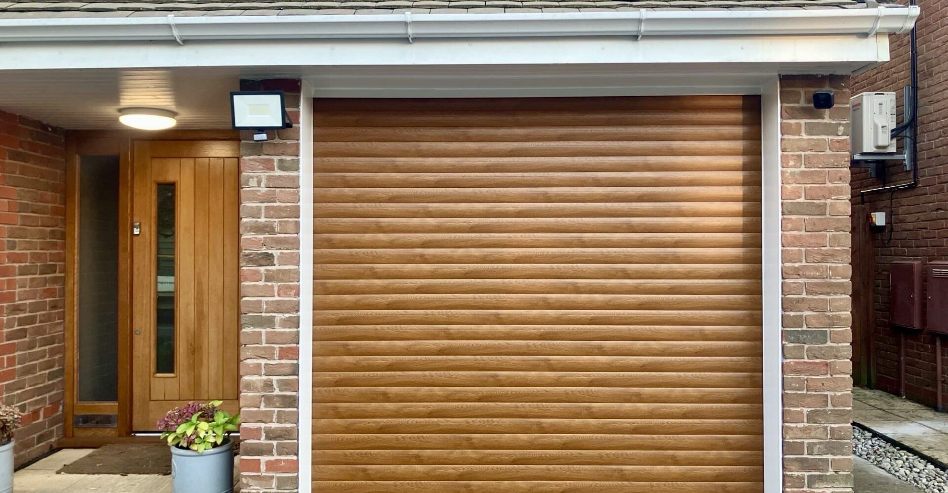 A brown roller garage door with horizontal slats, adjacent to a wooden front door and brick wall.