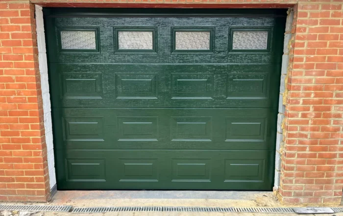 Green garage door with rectangular windows, set within a brick wall.